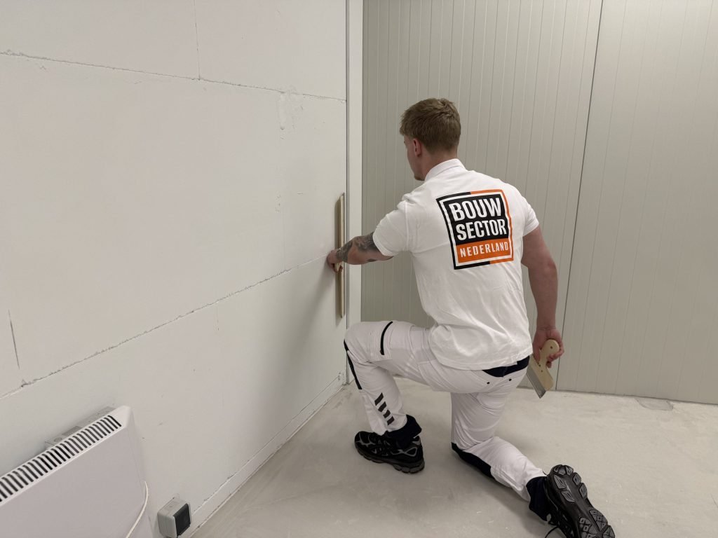 Stukadoor zit geknield bij een wand van gipsblokken en werkt met de spaan de onderkant bij de vloer af